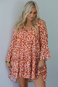 A woman wearing an orange and white animal print long sleeved, flowy mini dress, standing indoors against a white wall.