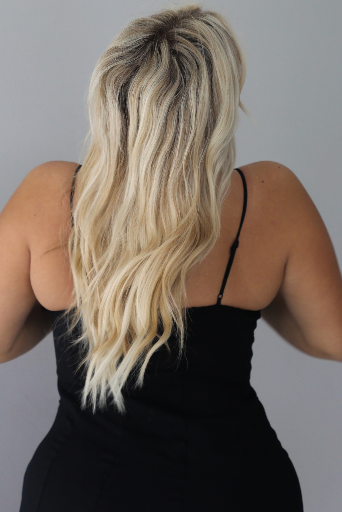 A woman shown from behind wearing a black formal maxi dress with lace trim detailing at the bodice and ruffled details at the skirt, standing indoors against a white wall. The back shows the adjustable straps.