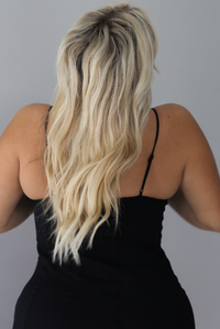 A woman shown from behind wearing a black formal maxi dress with lace trim detailing at the bodice and ruffled details at the skirt, standing indoors against a white wall. The back shows the adjustable straps.