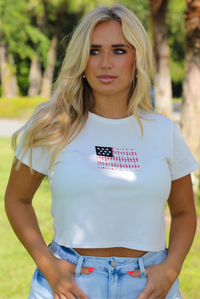 Woman wearing a white baby tee with an embroidered American Flag on the front with light wash denim shorts outdoors with greenery and palm trees in the background. 