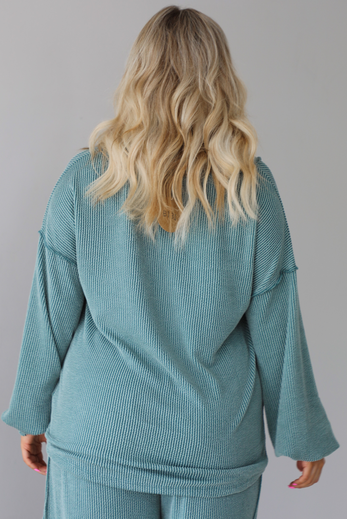 Woman wearing a teal two piece set that has a v-neck long sleeved oversized top and high waisted pants. The top is untucked to show its full length. The photo shows the back of the top. The photo is taken indoors against a white wall.