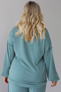 Woman wearing a teal two piece set that has a v-neck long sleeved oversized top and high waisted pants. The top is untucked to show its full length. The photo shows the back of the top. The photo is taken indoors against a white wall.