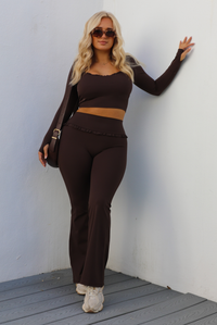 Woman model wearing a brown two-piece athleisure set. The top is cropped and the pants are high waisted. The photo is taken outdoors against a white wall.