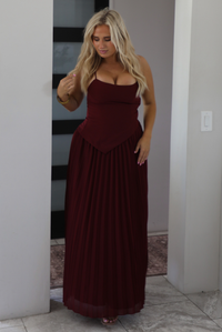 A woman wearing a wine formal maxi dress standing indoors in a modern home in an archway with a black and white opaque paneled door in the background. 