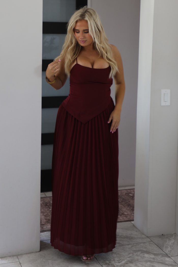 A woman wearing a wine formal maxi dress standing indoors in a modern home in an archway with a black and white opaque paneled door in the background. 