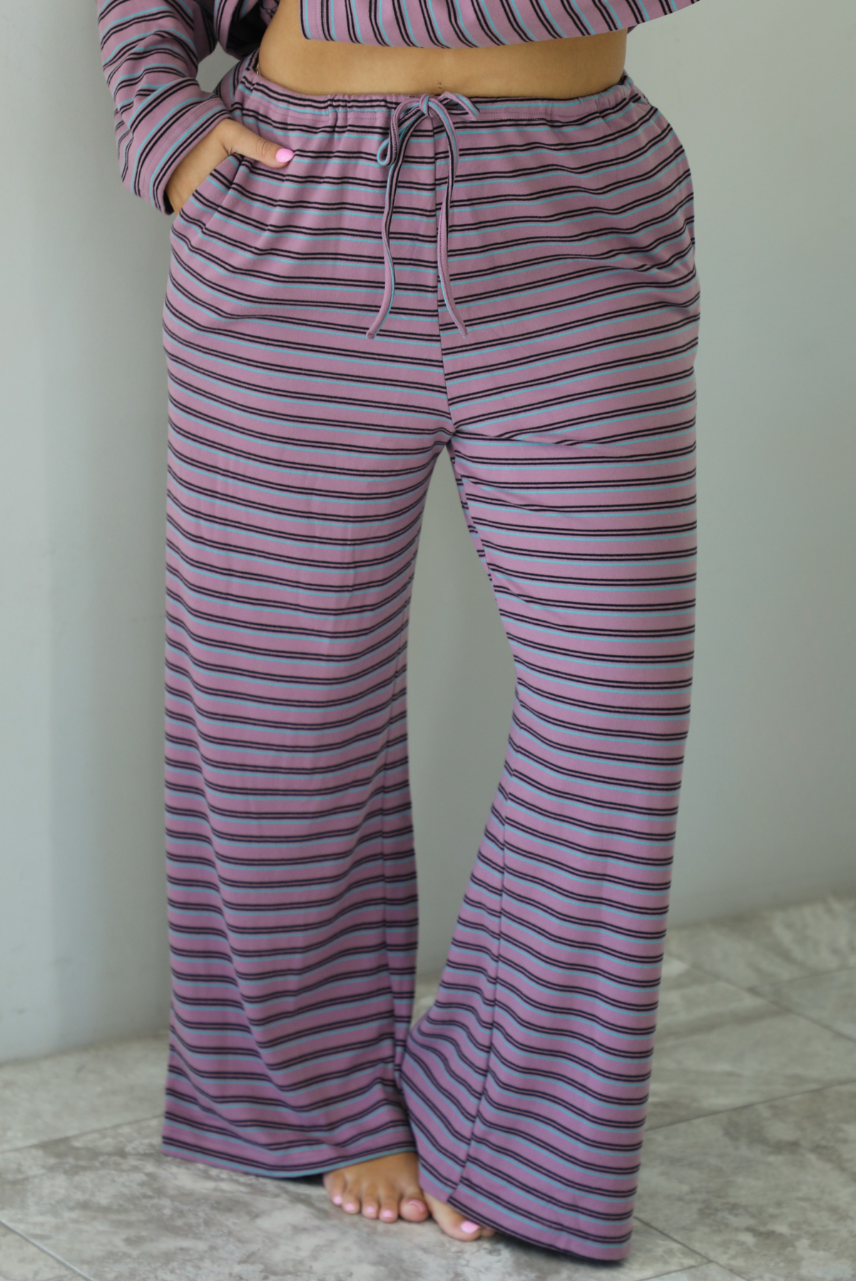 A woman shown from the waist down wearing a purple matching set with a black and blue striped pattern featuring an oversized long sleeved top and high waisted flared hem pants, standing indoors against a white wall with white marble flooring. The photo shows a closeup of the pants, which have an elastic and drawstring waistline with functional pockets on either side. 