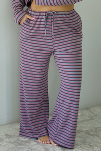 A woman shown from the waist down wearing a purple matching set with a black and blue striped pattern featuring an oversized long sleeved top and high waisted flared hem pants, standing indoors against a white wall with white marble flooring. The photo shows a closeup of the pants, which have an elastic and drawstring waistline with functional pockets on either side. 