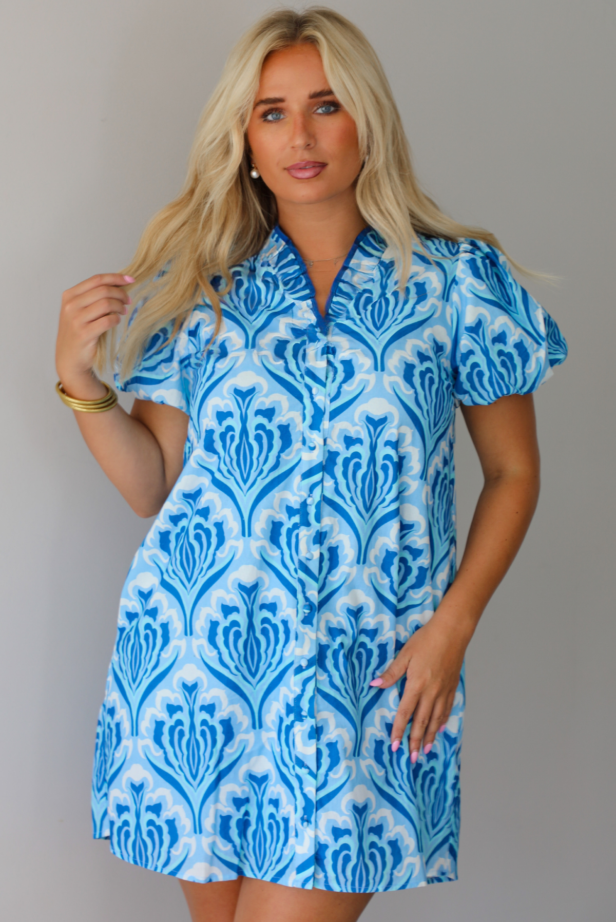 A woman modeling a short-sleeve button-up mini dress with a blue and white pattern, standing indoors against a white wall.