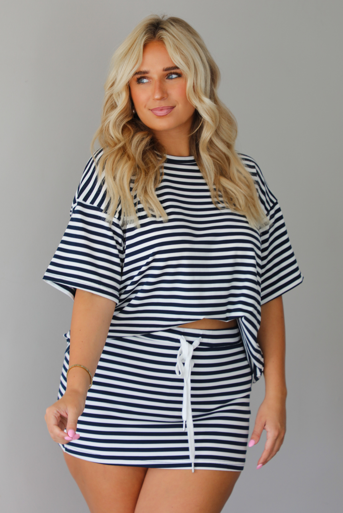 A woman wearing a navy and white horizontally striped short-sleeve oversized tee and skirt with a white drawstring tie at the waist, standing indoors against a white wall.