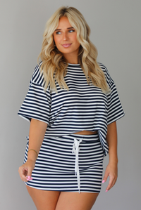 A woman wearing a navy and white horizontally striped short-sleeve oversized tee and skirt with a white drawstring tie at the waist, standing indoors against a white wall.