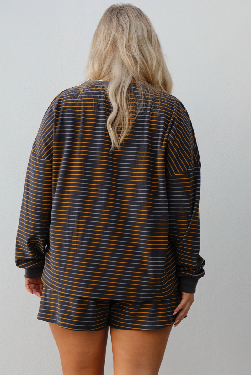 Close up of the back of a woman model wearing a brown and multi-colored striped pattern throughout. The set consists of an oversized top with quarter-button details and shorts. The photo is taken outdoors against a white wall. The photo showcases the full length of the top that stops mid-hip on our 5'7" model.