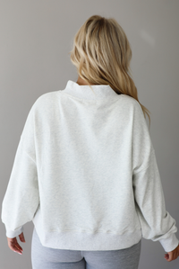 A woman shown from behind posing in a light gray sweatshirt with 'AMOUR' printed in gray letters and light heather gray leggings, standing indoors against a white wall. The photo shows a closeup of the back of the sweatshirt, which is devoid of any designs or graphics. 
