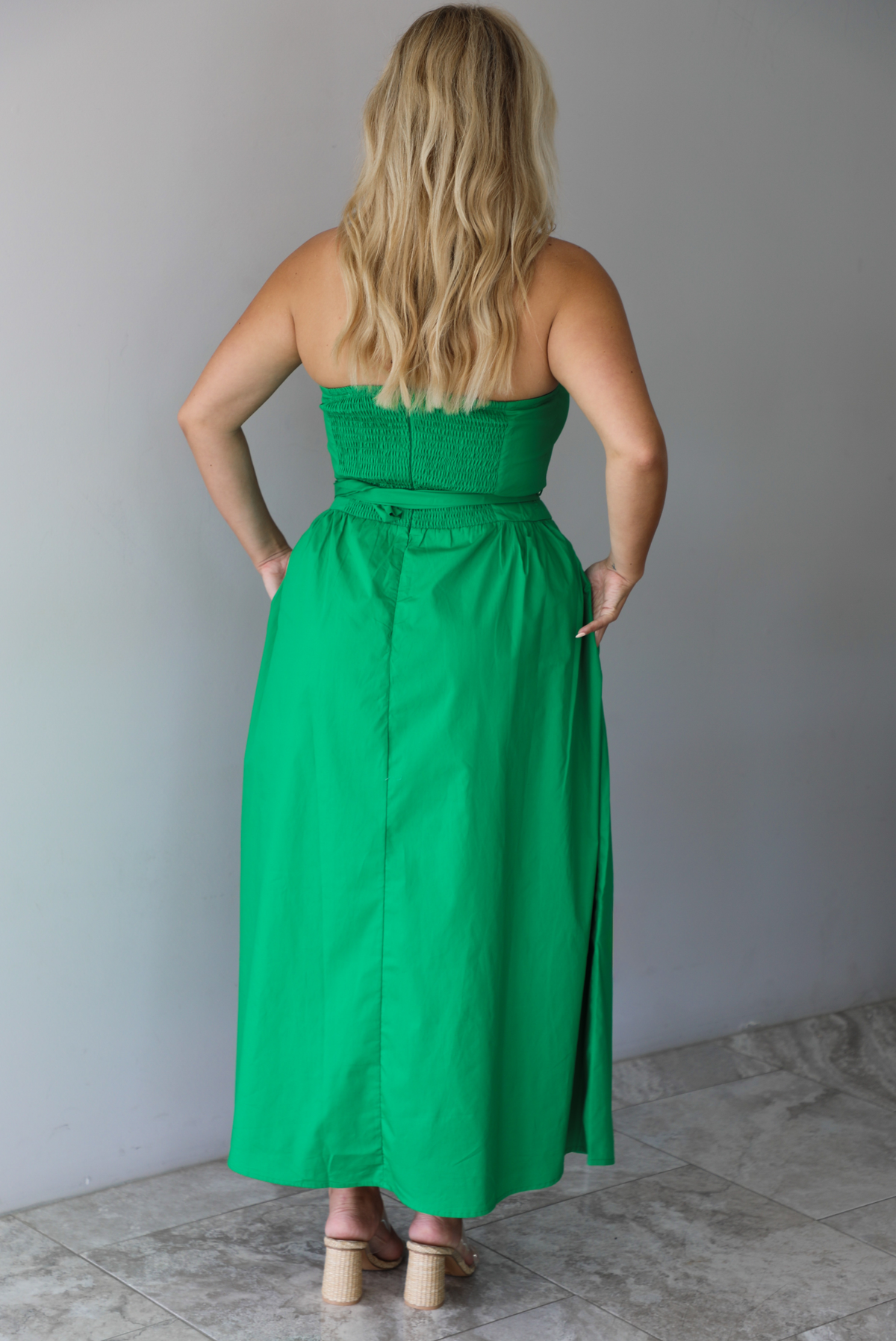 Woman wearing a green strapless midi dress that has a fitted bodice, belted waistline detail, functional pockets on either side, and a midi length indoors against a white wall with white marble flooring. The photo shows the back of the dress, which is smocked, and has a zipper closure. 