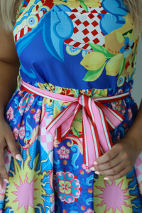 Woman wearing a multi-colored and multi-patterned mini dress with thick red straps, a cinched waistline with a removable tie, and a flowy skirt indoors against a white wall. The photo shows a closeup of the waistline of the dress. 