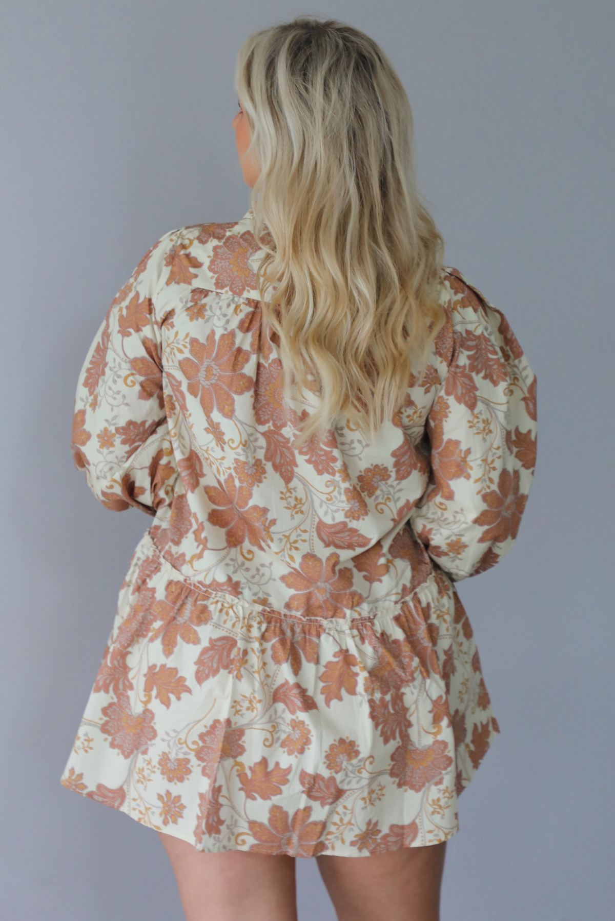 A woman shown from behind wearing a cream dress with a tan fall floral print pattern throughout, collared neckline, functional button closures along the front, long sleeves that cinch tighter at the wrist, and a slightly flowy skirt, standing indoors against a white wall.