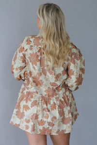 A woman shown from behind wearing a cream dress with a tan fall floral print pattern throughout, collared neckline, functional button closures along the front, long sleeves that cinch tighter at the wrist, and a slightly flowy skirt, standing indoors against a white wall.