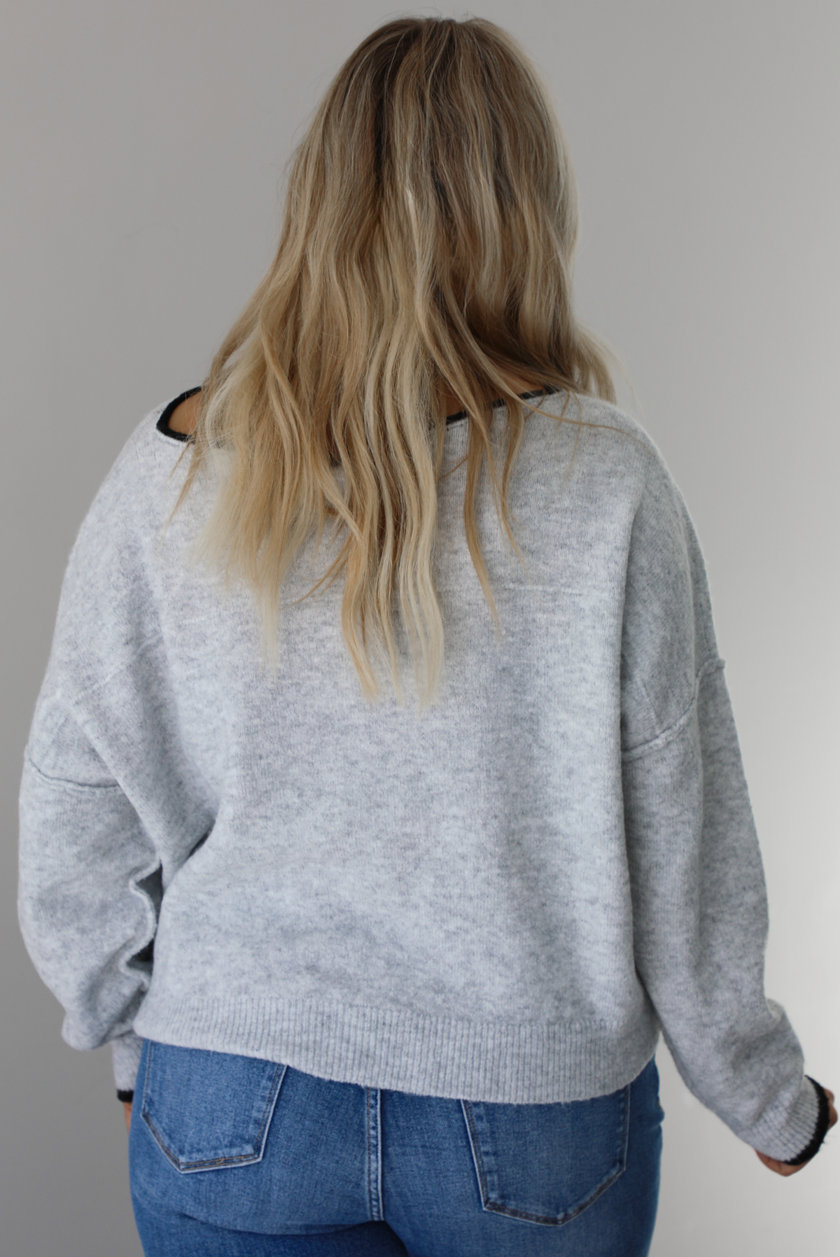 Woman wearing a grey relaxed fit sweater that has black trim on it, and medium wash distressed denim jeans. The photo shows the back of the sweater.  The photo is taken indoors against a white wall.