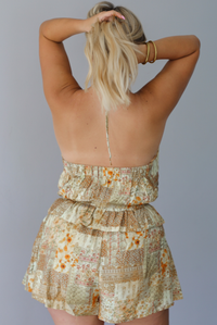 A woman shown from the back wearing a halter neckline yellow and multi patchwork patterned romper that has a cinched waistline, a ruffled detail at the waistline, and functional pockets on either side, standing indoors against a white wall. 