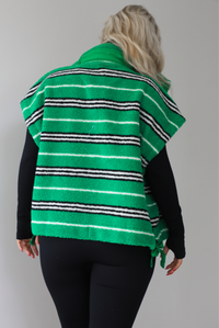 Woman wearing a green sherpa vest with black and white stripes that has an oversized fit, with a long sleeved, fitted, full body jumpsuit. The photo shows a close up of the back of the vest. The photo is taken indoors against a white wall with white marbled flooring. 