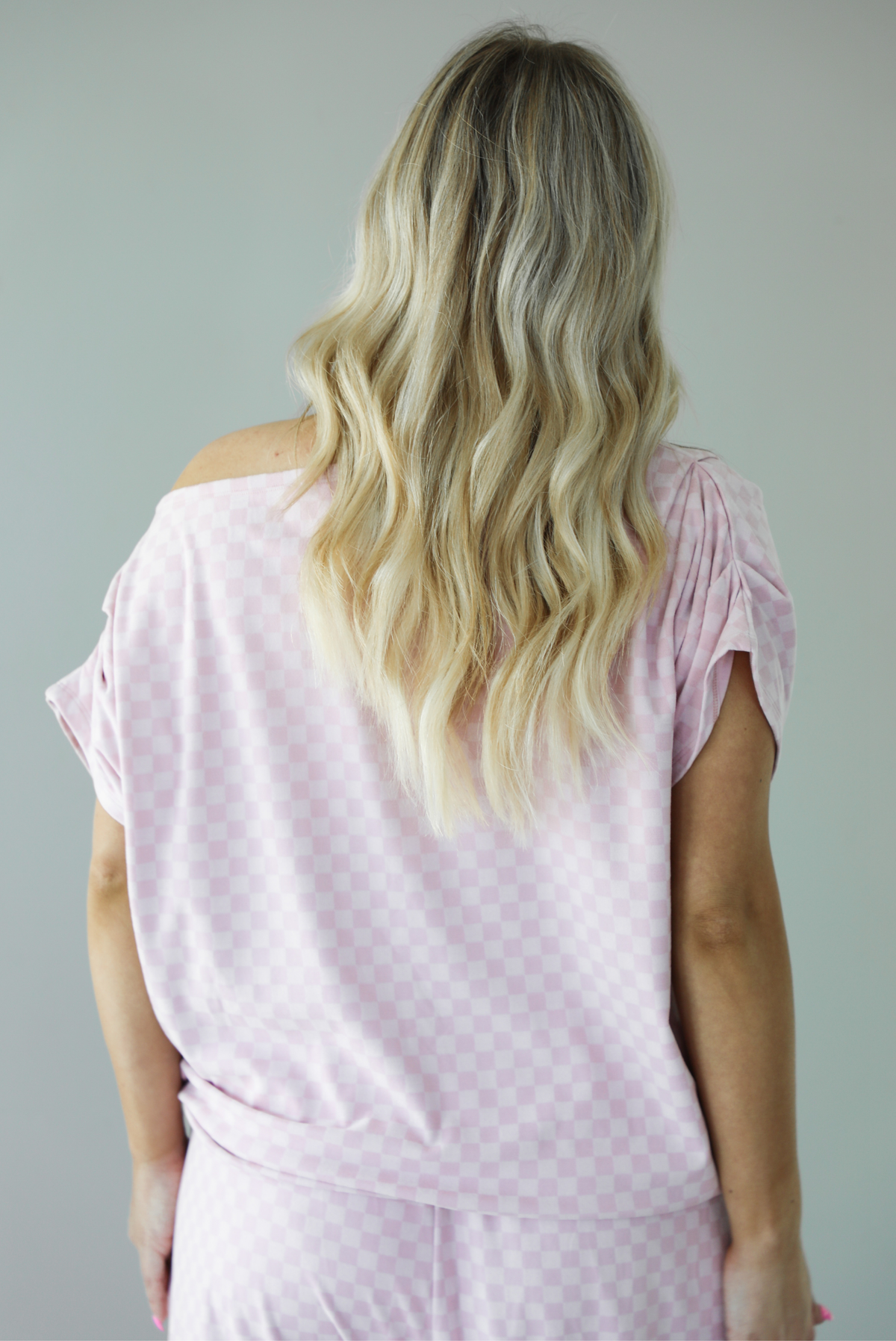 Woman wearing a pink checkered set featuring a slouchy top and pants. The photo is taken indoors against a white wall and shows a closeup of the back of the top, which is worn off the shoulders and has a relaxed fit, and is untucked to show the full length. 