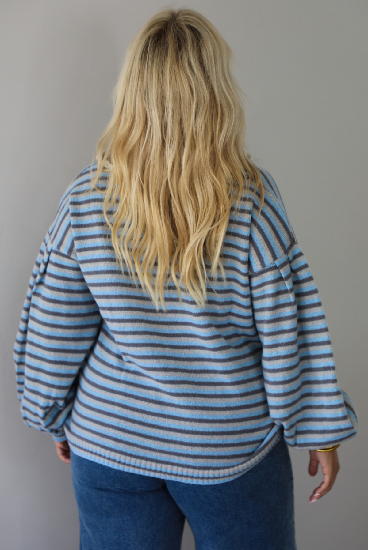 A woman shon from behind wearing a blue and navy and light blue striped knit sweater with a white lace collar, paired with blue jeans, standing against a plain white wall. 