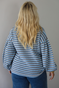 A woman shon from behind wearing a blue and navy and light blue striped knit sweater with a white lace collar, paired with blue jeans, standing against a plain white wall. 