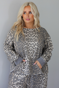 A woman wearing a leopard patterned two piece set featuring an oversized top with a front pocket detail and mid-rise oversized pants, standing indoors against a white wall. The photo shows a closeup of the top, showing the models hands in the front pocket. 
