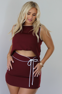 A woman in a two-piece burgundy set consisting of a sleeveless top that's bra tucked giving it a cropped look and a matching mini skirt with white trim detailing, and a tie waist detail, paired with black pointed-toe heels, standing indoors against a white wall.