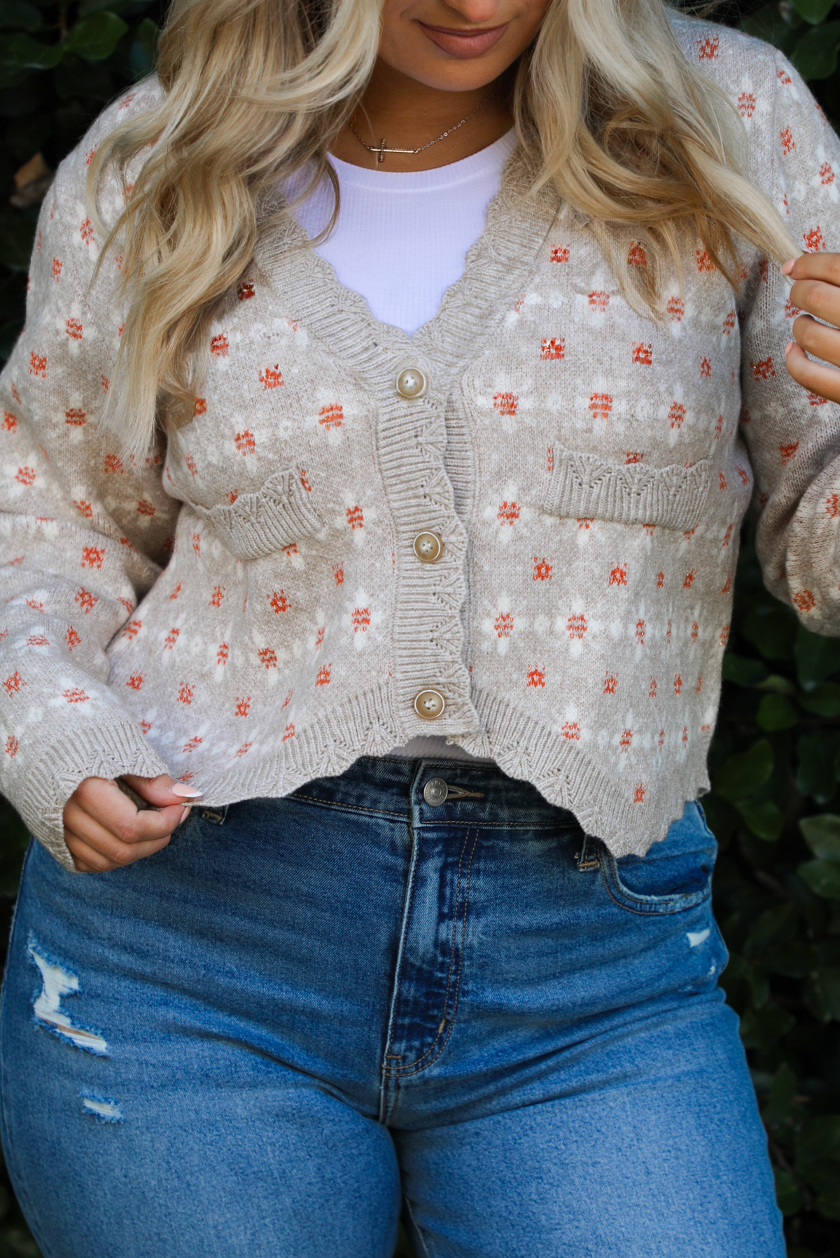 Woman model wearing a grey cardigan that has an orange and white floral print pattern throughout, long sleeves, an asymmetrical hem, and functional button closures in the front, with a long sleeved white top, and a pair of medium wash denim jeans outdoors in front of a green hedge. The photo shows a close up of the cardigan and the scalloped hemline, functional button closures in the front, and the faux-pockets in the front. 