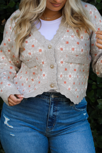 Woman model wearing a grey cardigan that has an orange and white floral print pattern throughout, long sleeves, an asymmetrical hem, and functional button closures in the front, with a long sleeved white top, and a pair of medium wash denim jeans outdoors in front of a green hedge. The photo shows a close up of the cardigan and the scalloped hemline, functional button closures in the front, and the faux-pockets in the front. 