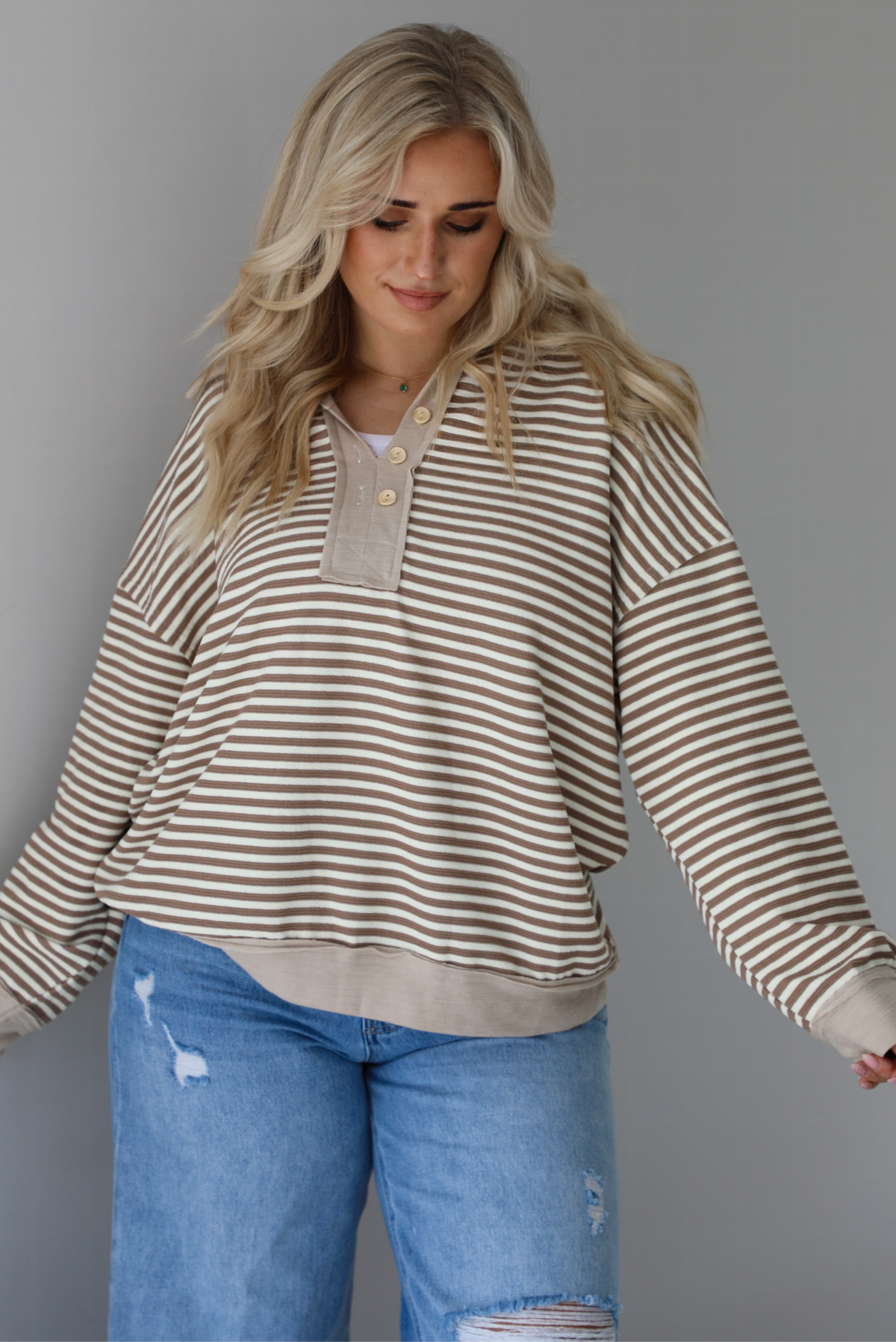 Woman wearing a mocha and cream striped pullover with a collared neckline, quarter button closures in the front, that has an oversized fit. The photo is taken indoors against a white wall.