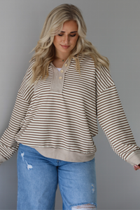 Woman wearing a mocha and cream striped pullover with a collared neckline, quarter button closures in the front, that has an oversized fit. The photo is taken indoors against a white wall.