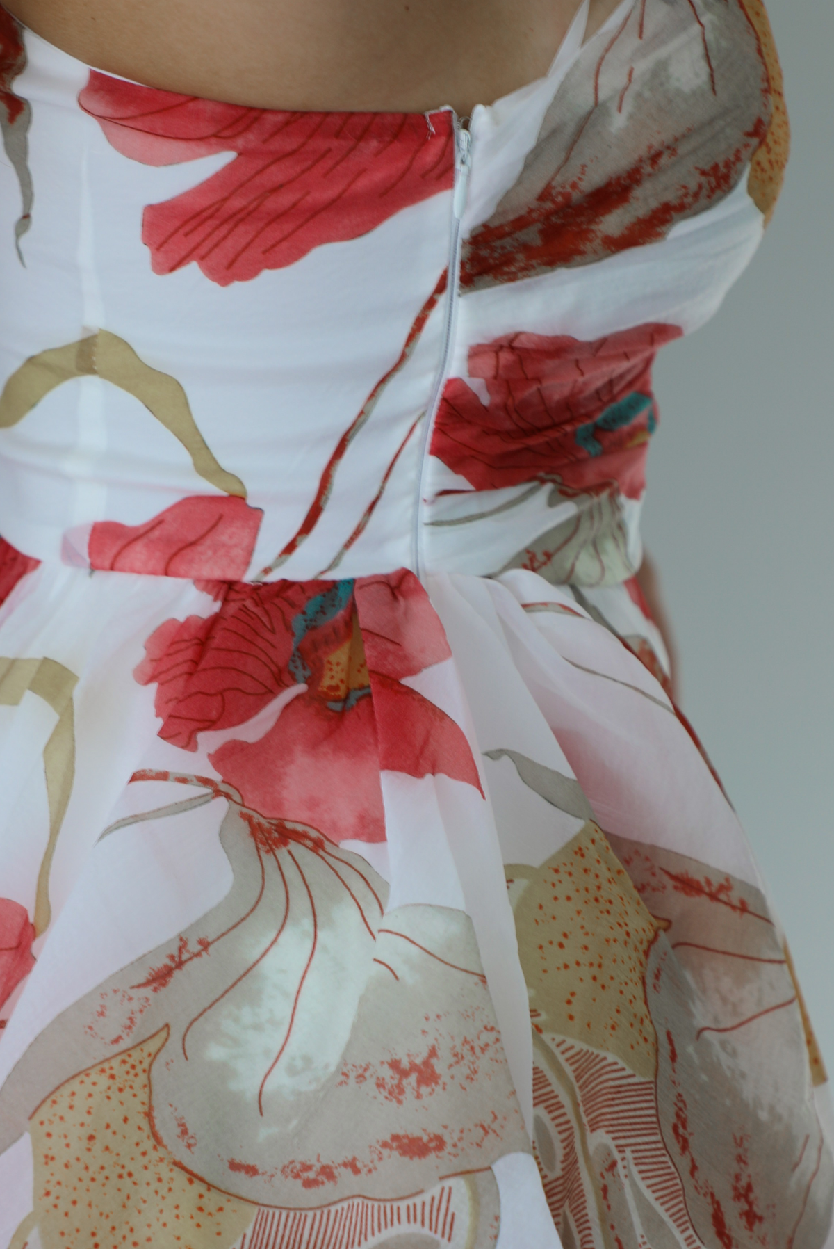 Woman wearing a one strap white and multi-colored floral dress with a ruffled skirt. The photo is taken indoors against a white wall and shows the zipper closure on the right side. 
