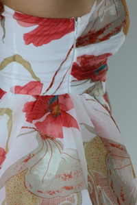 Woman wearing a one strap white and multi-colored floral dress with a ruffled skirt. The photo is taken indoors against a white wall and shows the zipper closure on the right side. 