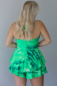Woman waring a green and multi-colored halter neck romper that has cut out details under the bust with a ruffled skirt design that makes it look like a dress indoors against a white wall. It shows the back of the romper, which has a zipper closure.