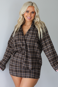 A woman wearing aa dark brown plaid set featuring an oversized button-down and fitted mini length skirt, with one side of the top front tucked into the skirt, standing indoors against a white wall.