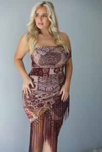 A woman wearing a burgundy and multi-colored patterned dress with a faux-wrap design, off the shoulder one sided sleeve, and fringe detailing at the hem, standing indoors against a white wall.