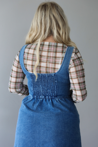 A woman shown from behind wearing a medium wash denim midi dress with button closures along the  front, with pockets on either side, paired with a sheer, multi-colored plaid long sleeved top underneath, standing indoors against a white wall with white marble flooring. The back shows a smocked detail. 