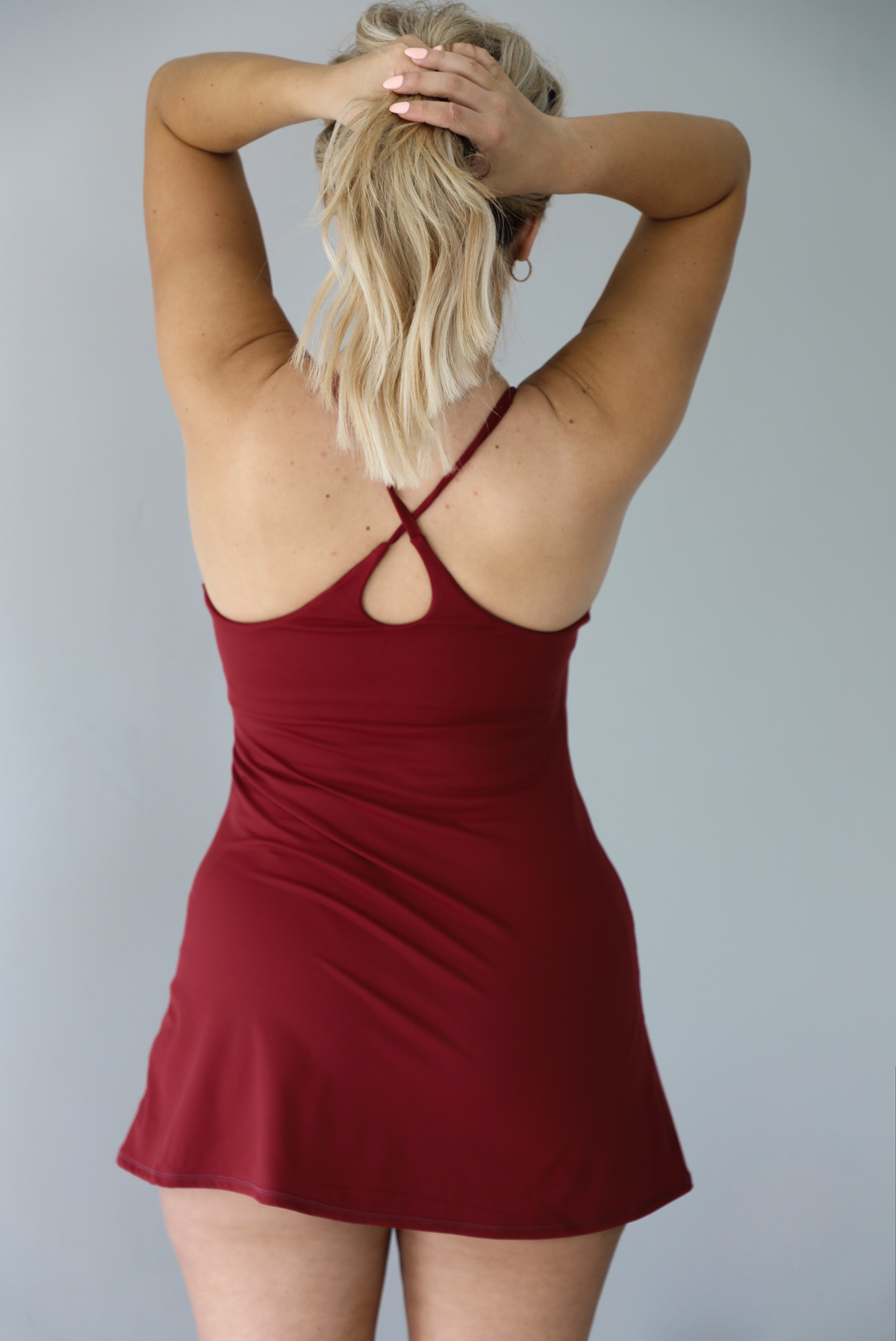 Woman wearing a maroon athleisure dress with thin straps and a relaxed fit indoors against a white wall. The photo shows the back of the dress, which the crisscross straps.