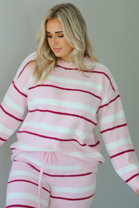 Woman wearing a pink set with a darker pink and white striped design. The set consists of an oversized sweater top and high waisted pants. The sweater of the set is tucked slightly in the front of the pants. The photo shows a closeup of the sweater. The photo is taken indoors against a white wall. 