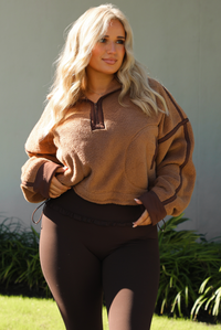 Woman model wearing a tan and brown quarter zip sweater with dark brown athleisure pants. The photo is taken outdoors against some greenery and a white wall. The photo showcases the sweater close up. 