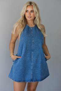 Woman wearing a denim blue dress indoors against a white wall. The photo shows a closeup of the dress, which has functional pockets on either side. 