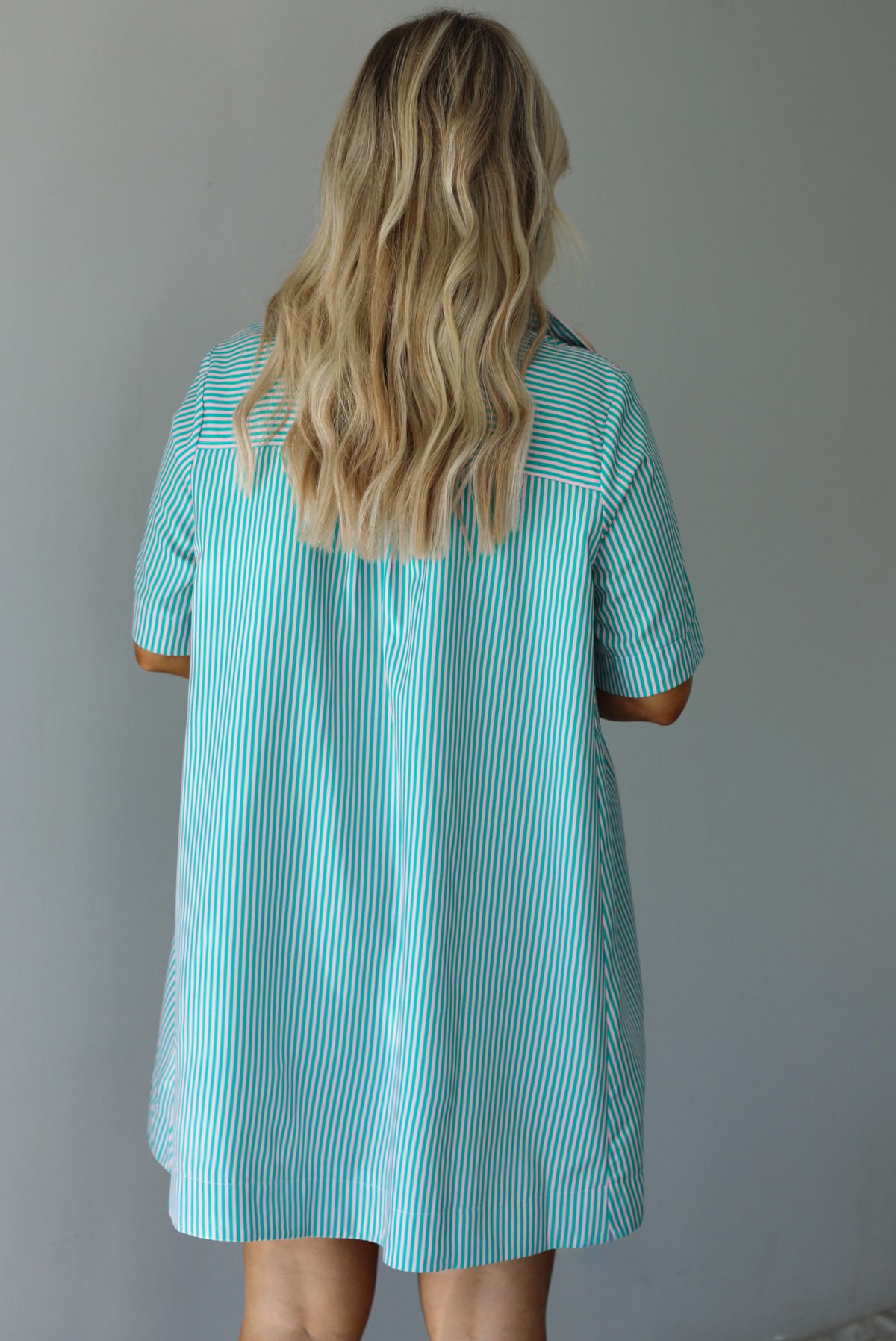 Woman wearing a green and pink striped dress that has a collared neckline, functional quarter button closures in the front, and a flowy skirt. The photo is taken indoors against a white wall and shows the back of the dress. 