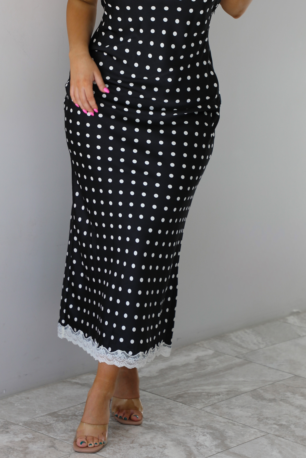 Woman wearing a black wearing a white polka dot printed midi dress that has a halter tie neckline, is form fitting, with white lace trim at the bottom. The photo is taken indoors against a white wall and shows a closeup of the skirt of the dress. 