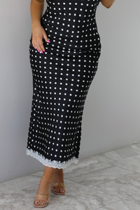 Woman wearing a black wearing a white polka dot printed midi dress that has a halter tie neckline, is form fitting, with white lace trim at the bottom. The photo is taken indoors against a white wall and shows a closeup of the skirt of the dress. 