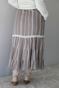 A closeup of a woman from behind wearing a taupe with white floral embroidered pattern maxi skirt, standing indoors against a white wall with white marble flooring. 
