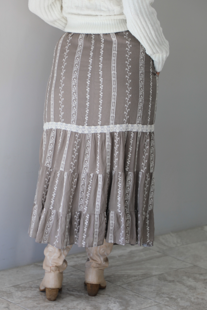 A closeup of a woman from behind wearing a taupe with white floral embroidered pattern maxi skirt, standing indoors against a white wall with white marble flooring. 