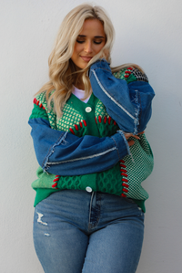 Woman model wearing a green and multi-colored cardigan with denim sleeves, and medium wash denim jeans. The photo shows up a close up of the cardigan and the stitched details, along with the frayed, raw hem details on the sleeves. The photo is taken against a white wall outdoors.