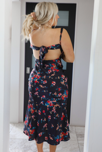 Back up of the top half of a woman model wearing a navy dress with a multi-colored floral print. The dress is a satin material, is a midi length, and is curve-hugging. The photo is taken inside a modern white and black interior. The photo showcases the adjustable straps, open back, and tie closure in the back.