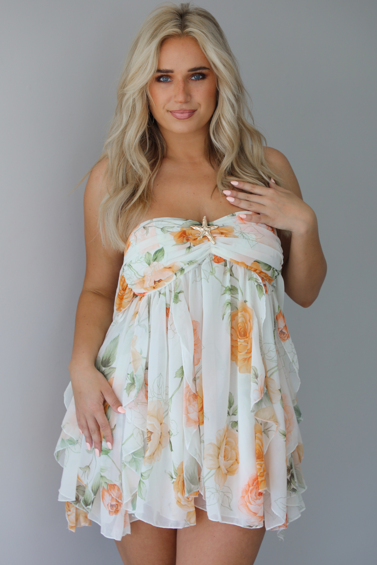 Woman wearing a white, strapless, babydoll cut dress that has an orange and green floral print pattern and flowy skirt indoors against a white wall.